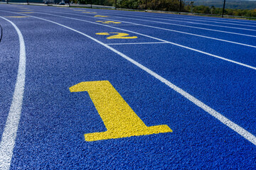 Close up photo of outdoor running track lane yellow number one, 1, on a new blue track with white lane lines.