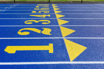 Close up photo of outdoor blue running track lanes with yellow, gold, numbers one, two, three, four five, six, seven, eight, 1, 2, 3, 4, 5, 6, 7, 8, with on a new track with white lane lines.