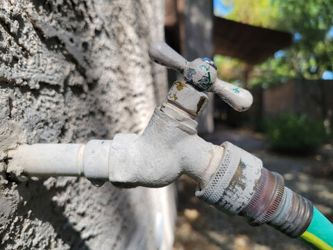 Close-up shot of an old, weathered traditional outdoor water faucet with backflow preventer attached to a textured, stucco wall in out of focus yard