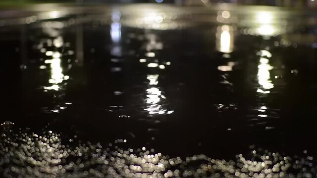 Night scene of rain puddle reflecting city lights on wet asphalt road surface, creating abstract light patterns and bokeh effect.