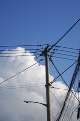 Fototapeta premium A utility pole with multiple wires and a street lamp attached, set against a clear blue sky with some white clouds.