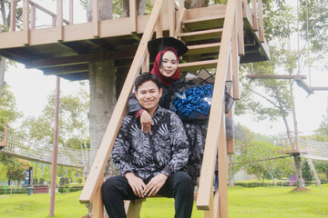 Indonesian couple in a happy moment and on a ladder from a tree house, with the man wearing typical Indonesian batik and the woman wearing an academic dress and decorated with a red hijab