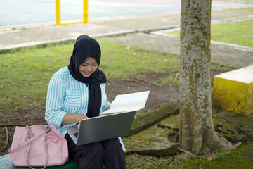 Young Muslim woman wearing a hijab diligently studying outdoors on a park bench with her laptop and books, embracing a modern lifestyle of remote learning and higher education