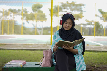 Serene young woman in a hijab and red headphones, deeply engrossed in reading a book while studying outdoors on a tranquil park bench, fostering knowledge and personal growth