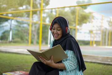 Serene young woman in a hijab and red headphones, deeply engrossed in reading a book while studying outdoors on a tranquil park bench, fostering knowledge and personal growth
