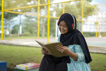 Serene young woman in a hijab and red headphones, deeply engrossed in reading a book while studying outdoors on a tranquil park bench, fostering knowledge and personal growth