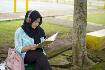 Serene young woman in a hijab and red headphones, deeply engrossed in reading a book while studying outdoors on a tranquil park bench, fostering knowledge and personal growth