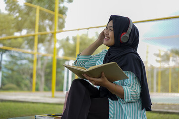 Serene young woman in a hijab and red headphones, deeply engrossed in reading a book while studying outdoors on a tranquil park bench, fostering knowledge and personal growth