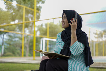 Serene young woman in a hijab and red headphones, deeply engrossed in reading a book while studying outdoors on a tranquil park bench, fostering knowledge and personal growth
