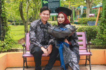 Indonesian couple in a happy moment in a park with the man wearing traditional Indonesian batik and the woman wearing an academic dress and decorated with a red hijab