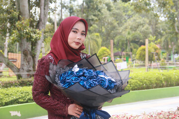 Beautiful Indonesian Muslim woman wearing a red hijab and holding a bouquet of blue flowers in a park filled with trees