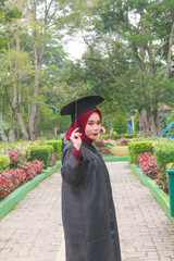 Beautiful Indonesian Muslim woman wearing maroon hijab and toga and academy dress appears elegant in a park