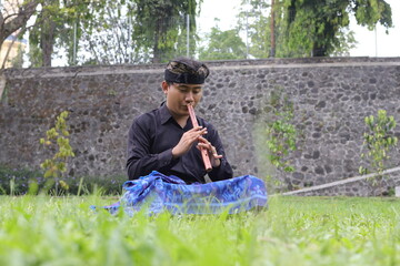 Outdoor shot of Asian man playing traditional bamboo flute in city park with green surroundings
