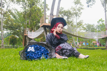 Beautiful Indonesian Muslim woman wearing maroon hijab and toga and academy dress sitting on the grass and accompanied by a blue bouquet of flowers