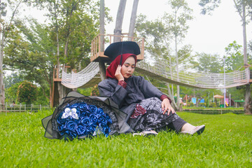 Beautiful Indonesian Muslim woman wearing maroon hijab and toga and academy dress sitting on the grass and accompanied by a blue bouquet of flowers