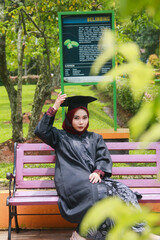 Beautiful Indonesian Muslim woman wearing maroon hijab and toga and academy dress sitting on a bench in a park