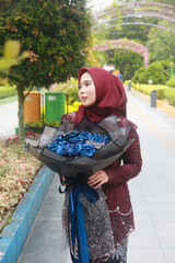 Beautiful Indonesian Muslim woman wearing a red hijab and holding a bouquet of blue flowers in a park filled with trees