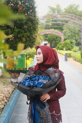Beautiful Indonesian Muslim woman wearing a red hijab and holding a bouquet of blue flowers in a park filled with trees