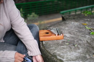Asian woman plays a kalimba in a green park on a sunny day. The calm melodies and lush leaves create a peaceful and relaxing scene