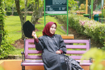 Beautiful Indonesian Muslim woman wearing maroon hijab and toga and academy dress sitting on a bench in a park