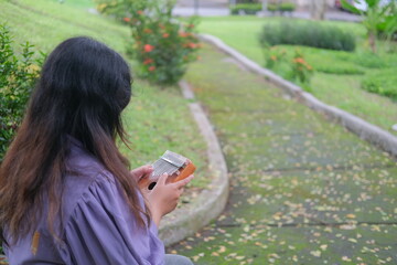 Asian woman relaxes in a city park on a sunny summer day, playing a traditional kalimba. Surrounded by greenery, she enjoys a peaceful moment, blending music with the tranquility of nature