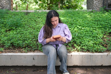 young Asian woman plays a kalimba while relaxing in a green city park during summer. The bright sunlight, lush surroundings, and soft melodies create a calm and refreshing outdoor scene