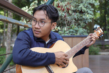 Portrait of Asian man playing guitar in a park during summer, surrounded by lush greenery and the peaceful atmosphere of the season
