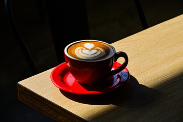 Close up view of hot cafe latte on the corner wooden table. red cup or mug. art. brown. dark, black, shadows. morning. relax, coffee time. cafe. coffee shop. sun shine, light. ceramic.
