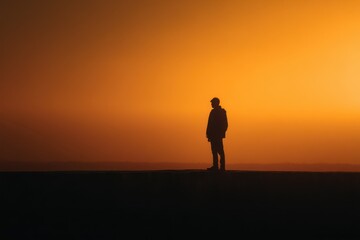 Fototapeta premium Silhouette of man standing alone at sunset with warm orange glow filling the sky, representing solitude, reflection, and peaceful connection with nature in minimal evening landscape