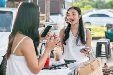 Pretty asian woman holding phone and talking with friend while sitting at food table in local market
