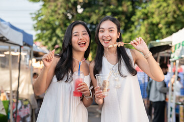 Pretty asian woman holding drink cup aside a friend eating street food as standing in local market.