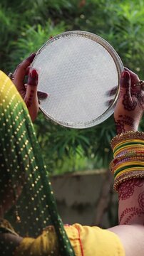 Indian married woman performing Karva Chauth ritual, holding a sieve to see the moon, wearing traditional saree, colorful bangles, and mehndi on hands during festival night.
