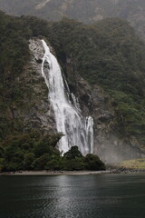 A powerful waterfall cascades down a sheer rock face into the dark waters of Milford Sound...