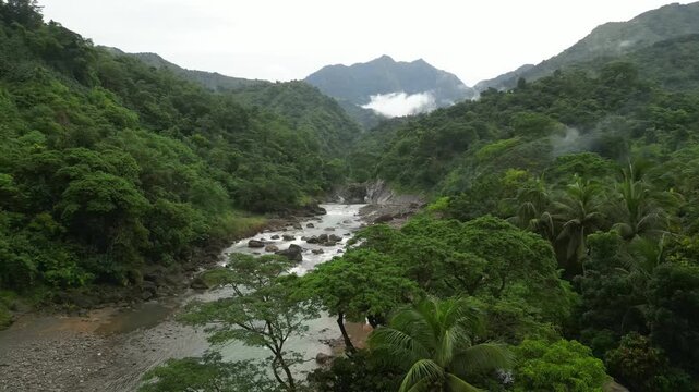 A wide aerial angle drifts to a final hold over a winding river and dense tropical forest. Mist clings to the mountain peaks as boulders and palm trees settle into the layered green valley.