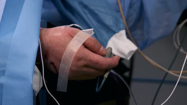 The hand of a patient with an IV undergoing surgery is seen from under a blue drape.