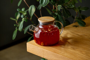A glass teapot filled with vibrant herbal tea sits on a smooth wooden table. Soft sunlight illuminates the scene, highlighting the rich colors and nearby green leaves, creating a calming atmosphere