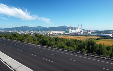Highway road extending towards an industrial complex with factory buildings, smokestacks, mountains, and a clear blue sky