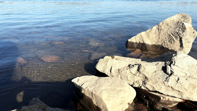 Coastal rocks in shallow water, large light-colored boulders at the edge of clear water, pebbles and sun-dappled ripples visible on the bottom. A minimalist natural backdrop with a sense of purity and