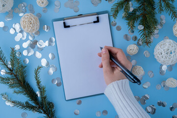 Female hands in white cozy sweater writing on empty white paper notepad on blue background. Preparation for New Year or Christmas. Copy space mock up Concept of planning and setting goals for personal