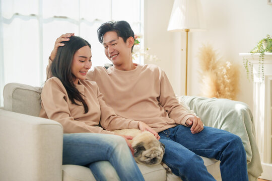 Happy Asian couple sitting close together on sofa, smiling and talking in a warm home atmosphere full of love and relaxation.
