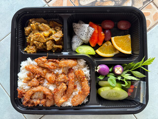 A typical Thai street food lunch box contains jasmine rice, fried pork, pork curry with vegetables, and fruit.