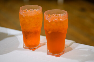 Two Refreshing Orange Colored Cold Drinks with Ice Cubes and Salt Rims on a White Tablecloth with a Blurry Wooden Background