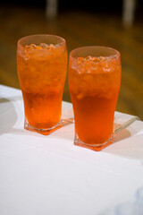 Two refreshing glasses of vibrant orange colored cold drinks filled with ice cubes and served on coasters on a white tablecloth perfect for summer parties or restaurant menus
