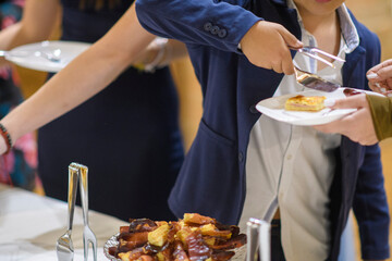 Young boy in blue blazer and white shirt carefully serves himself a pastry from a buffet table at a festive gathering occasion celebration event indoors