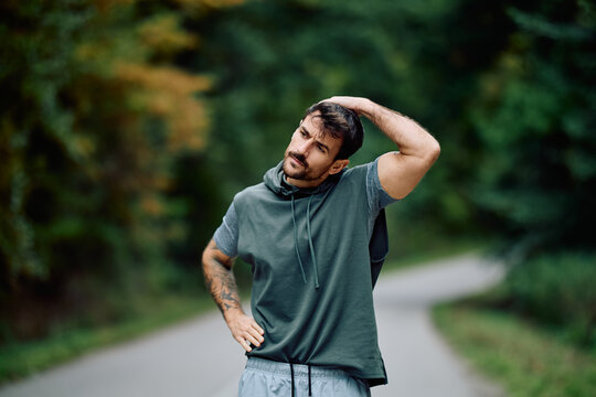 Athletic man in sportswear warming up by stretching neck muscles on a park road before an intense outdoor workout
