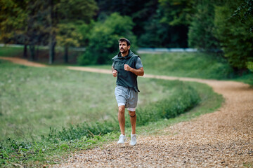 Young man jogging along a winding park path surrounded by green trees, focused on fitness,...