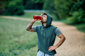 Man drinking red liquid from a bottle, resting during a fitness workout in a park, maintaining...