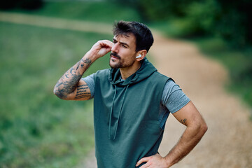 Athletic man adjusting a wireless earbud, enjoying music or a podcast while exercising outdoors in...