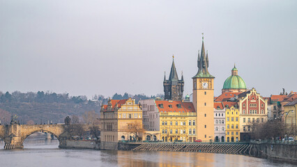 Panoramic view of Prague's Vltava River, featuring a historic bridge, colorful riverfront buildings, and a prominent clock tower under an overcast sky