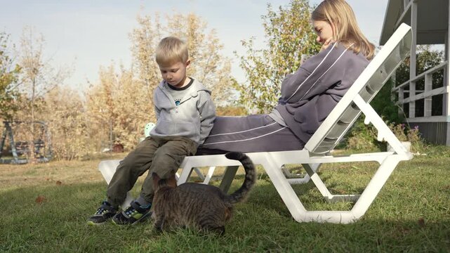 Two bored siblings, a boy and a girl, sitting on a deck chair in their backyard on a sunny day while a curious tabby cat sniffs around their feet, capturing a quiet moment of family life